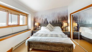 Een slaapkamer met een groot bed, houten hoofdeinde, twee nachtkastjes met lampen, een raam met zonwering en een muurschildering met bosmotief.