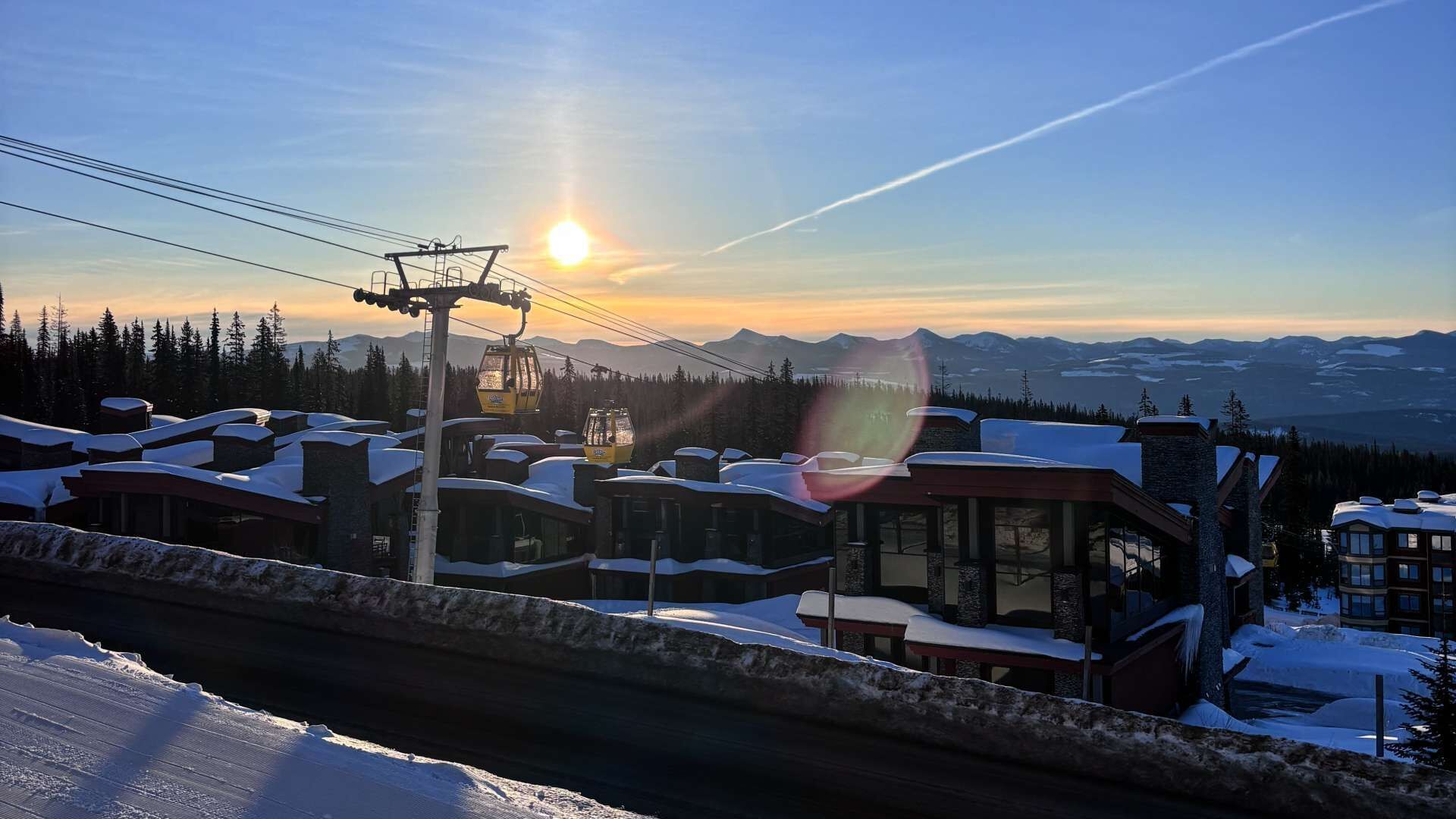 A snow-covered mountain resort with a cable car, cabins, and a view of the sun setting over the mountains.