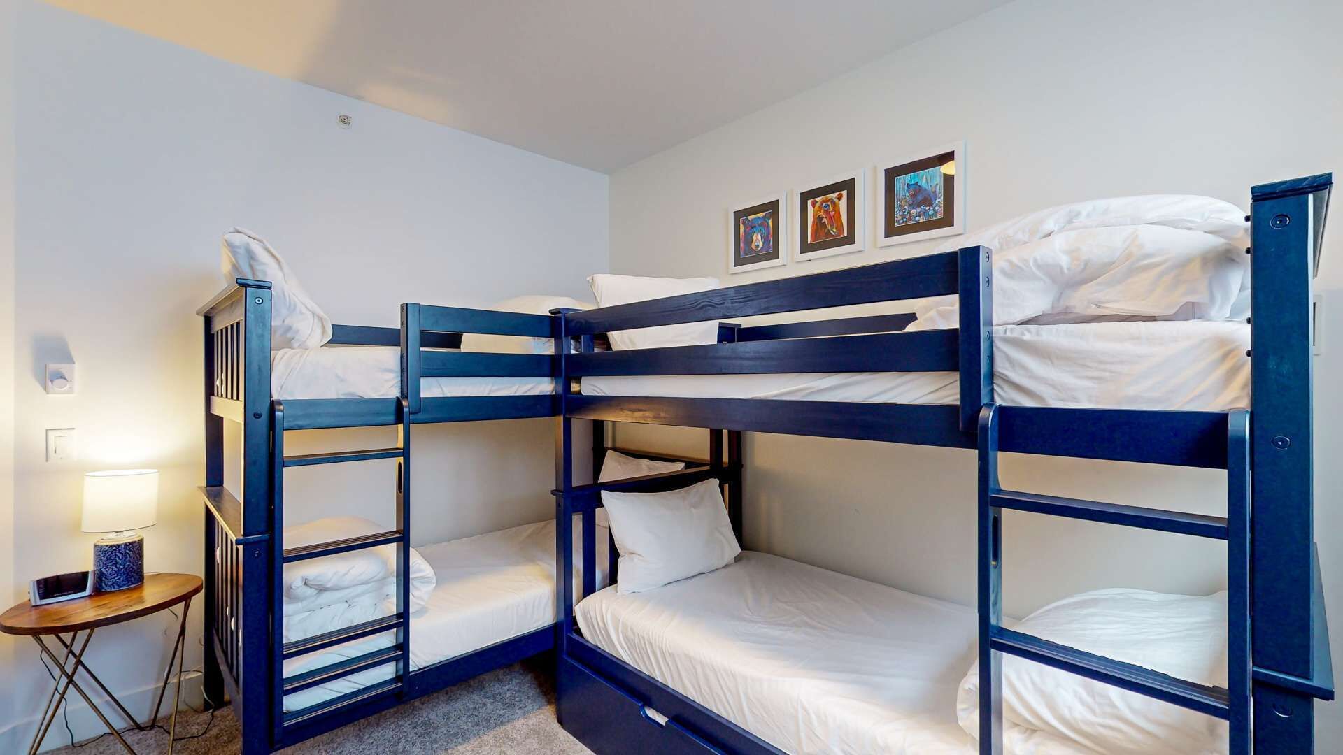 Habitación con literas, ropa de cama blanca, una mesita con lámpara y tres cuadros en la pared.