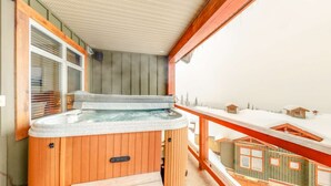Un jacuzzi en una terraza de madera con vista a cabañas nevadas.