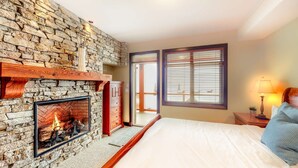Un dormitorio con chimenea de piedra, una cama con ropa de cama blanca, una mesita de noche con lámpara y una ventana con persianas.