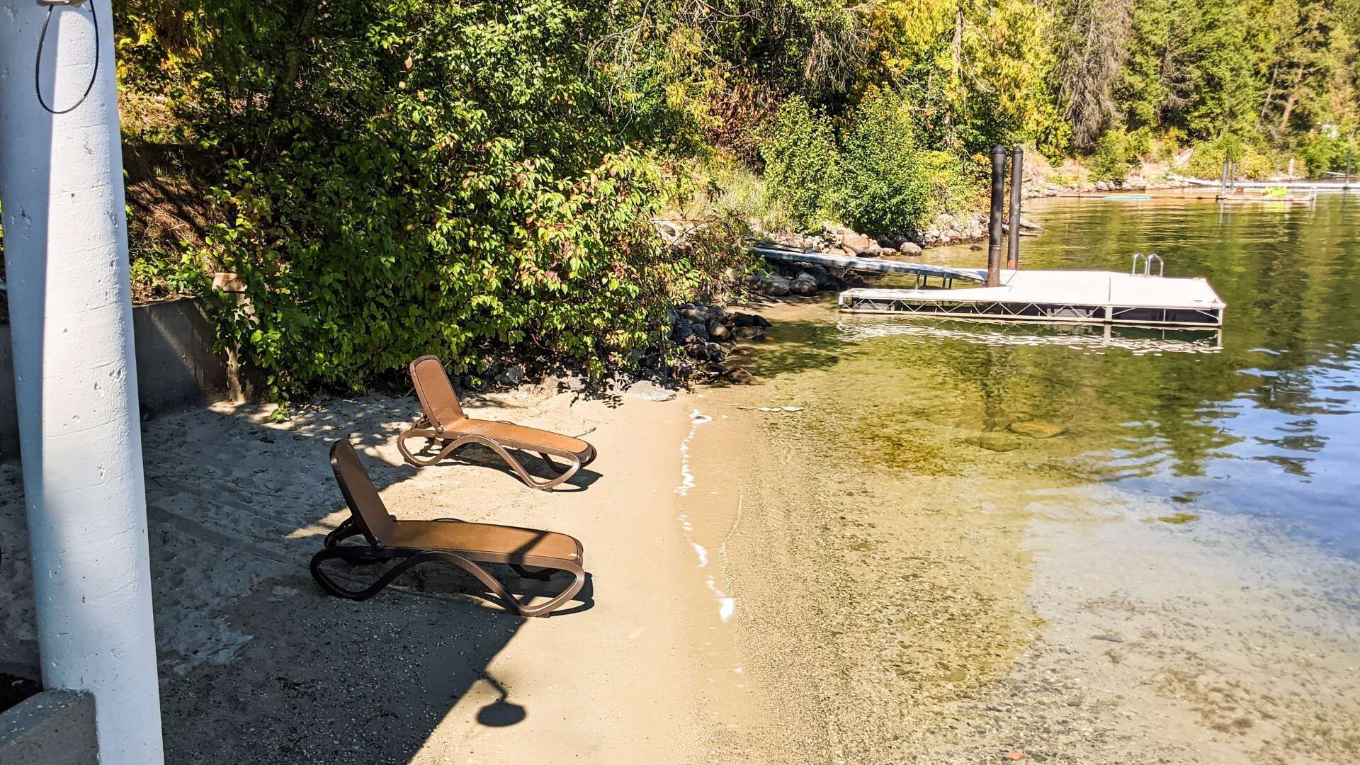 Une plage de sable avec deux chaises longues et une jetée qui s’avance dans un lac, le tout entouré d’une végétation luxuriante.