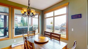 A dining area with a wooden table, chairs, and a chandelier. Large windows offer views of a ski lift and buildings. - Okanagan - Glacier Lodge 103 (Beaverdell)