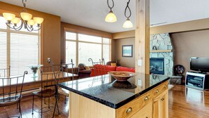 A spacious kitchen with a large island, wooden cabinets, and a stone fireplace. - Okanagan - Glacier Lodge 106 (Beaverdell)