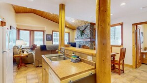 A cozy kitchen with a stone fireplace, wooden beams, and a large island with a sink. - Okanagan - Grizzly Lodge 306 (Beaverdell)