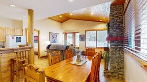 A spacious kitchen with wooden cabinets, a dining area with a large wooden table, and a stone accent wall. - Okanagan - Grizzly Lodge 306 (Beaverdell)