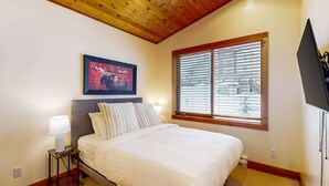 A bedroom with a bed, a bedside lamp, a framed picture of a moose, a window with blinds, and a TV on the wall. - Okanagan - Grizzly Lodge 306 (Beaverdell)