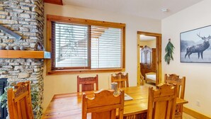 A dining room with a wooden table and chairs, a stone wall, and a large window with blinds. - Okanagan - Grizzly Lodge 306 (Beaverdell)