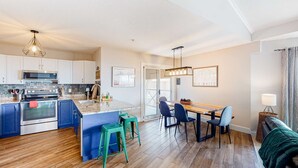A modern kitchen with a central island, a dining area with a wooden table and chairs, and a cozy seating area with a green sofa. - Okanagan - Hilltop Hideaway Condo - Legacy 401A (Beaverdell)