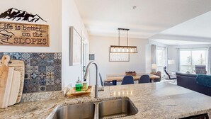 A modern kitchen with a granite countertop, stainless steel sink, and a wooden cutting board. There is a decorative sign on the wall and a dining area with a table and chairs. - Okanagan - Hilltop Hideaway Condo - Legacy 401A (Beaverdell)