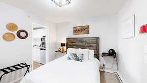 Quarto com cabeceira de madeira, cama com roupa de cama branca, mesa de cabeceira com abajur e relógio de parede.