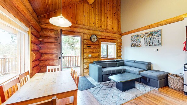 A log cabin interior with a wooden dining table, a gray sectional sofa, a clock, and a painting on the wall. - Okanagan - Lac Le Jeune at Cabin 4 (Kamloops)