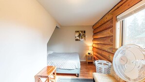 A cozy bedroom with a wooden log wall, a bed, a nightstand, a lamp, a fan, and a window with a view of trees. - Okanagan - Lac Le Jeune at Cabin 4 (Kamloops)