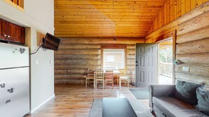 Interior de una cabaña de troncos con techo de madera, zona de comedor con mesa y sillas, un sofá gris y un refrigerador con un adhesivo de ardilla.