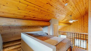 Un dormitorio de cabaña de madera con techo inclinado, cama con ropa de cama estampada, ventilador de techo y ventana con vistas.