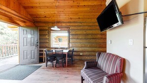 Interior de una cabaña de madera con comedor, sofá, televisor montado en la pared y puerta que da al exterior.