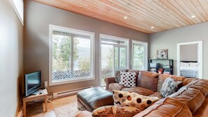 A living room with a brown leather couch, a TV on a wooden stand, and large windows with a view of trees. - Okanagan - IKES Retreat (Kootenay Boundary)