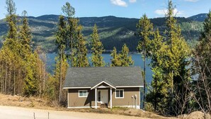 A single-story cabin with a gabled roof, a front porch, and a small deck. It is surrounded by tall trees and overlooks a lake with mountains in the background. - Okanagan - IKES Retreat (Kootenay Boundary)