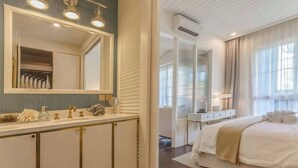 Moderni hotellihuone, jossa on suuri peili, meikkipöytä hygieniatuotteineen, beigen värinen peitto sängyllä ja näkymä käytävään.