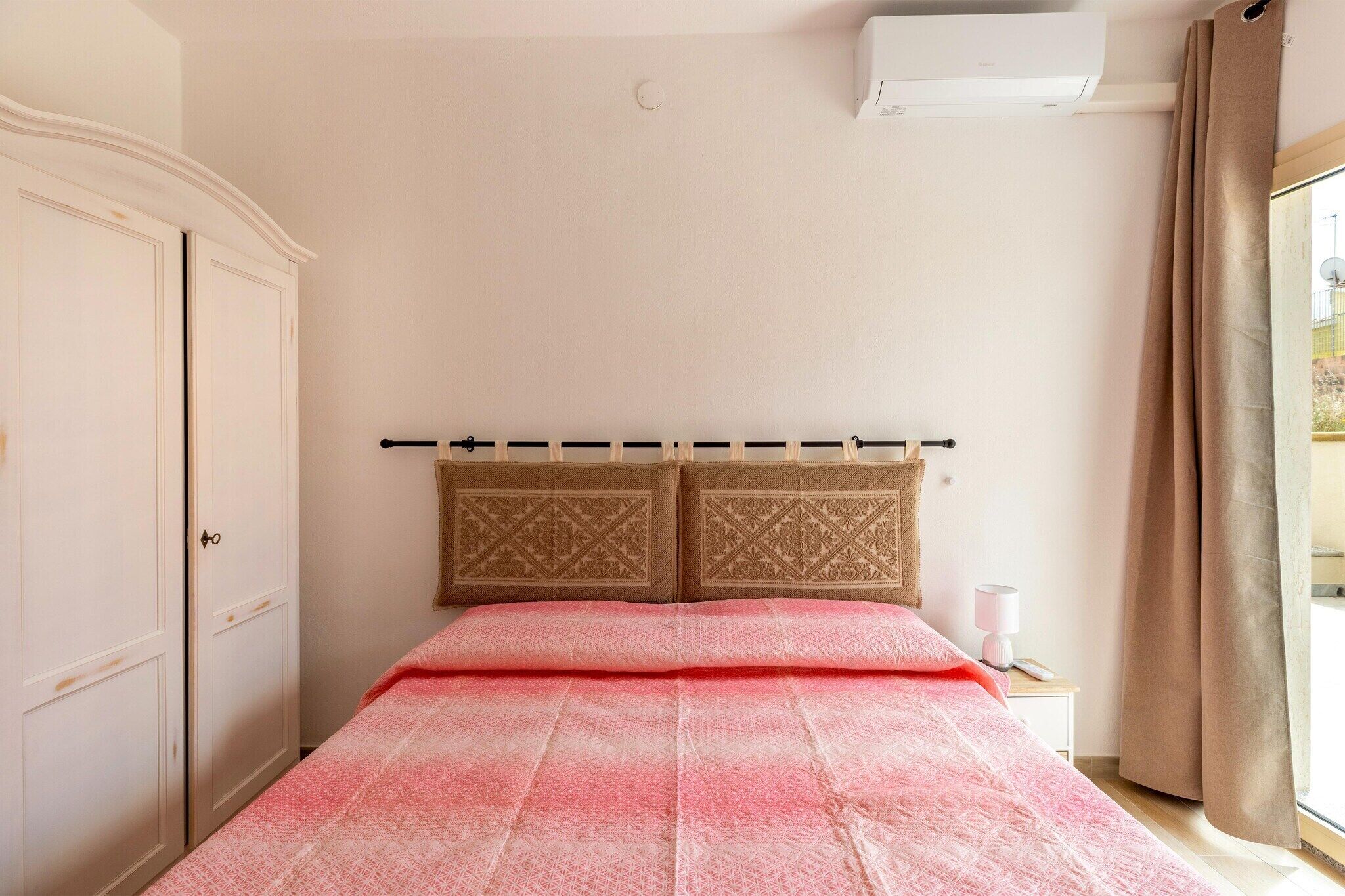 A bedroom with a bed, a wardrobe, a bedside table with a lamp, and a window with curtains.