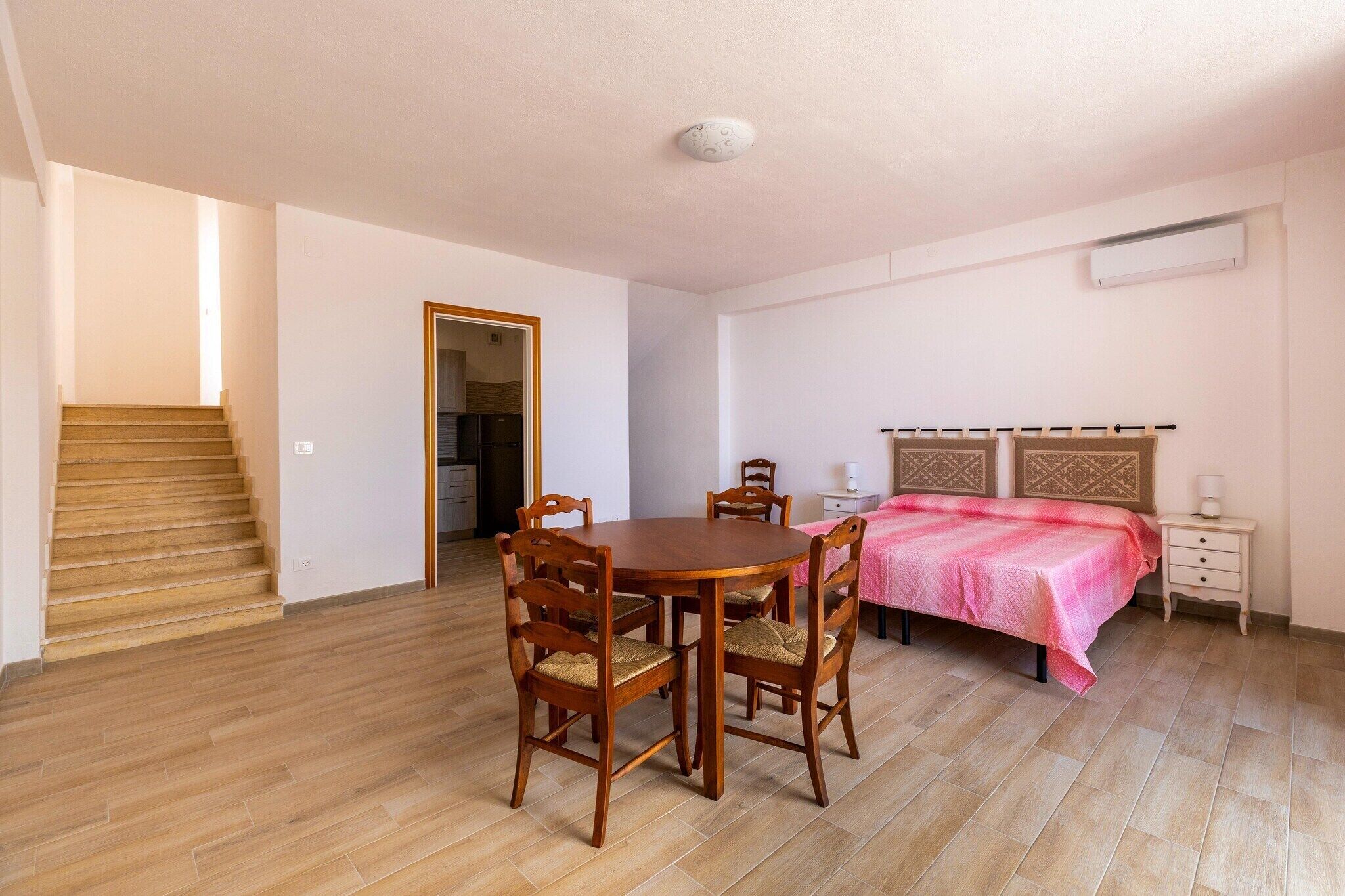 Una habitación con una mesa de comedor de madera, cuatro sillas, una cama con una manta rosa, una mesita de noche y una escalera que lleva a otra habitación.