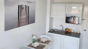 Een compacte keuken met witte kastjes, een magnetron en een spoelbak. Er is een eethoek met een tafel en stoelen, en een lijst met een afbeelding aan de muur.