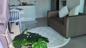 Een moderne woonkamer met een bruine bank, een witte salontafel en een grote groene plant. In de achtergrond is een keuken zichtbaar met witte kasten en een magnetron.
