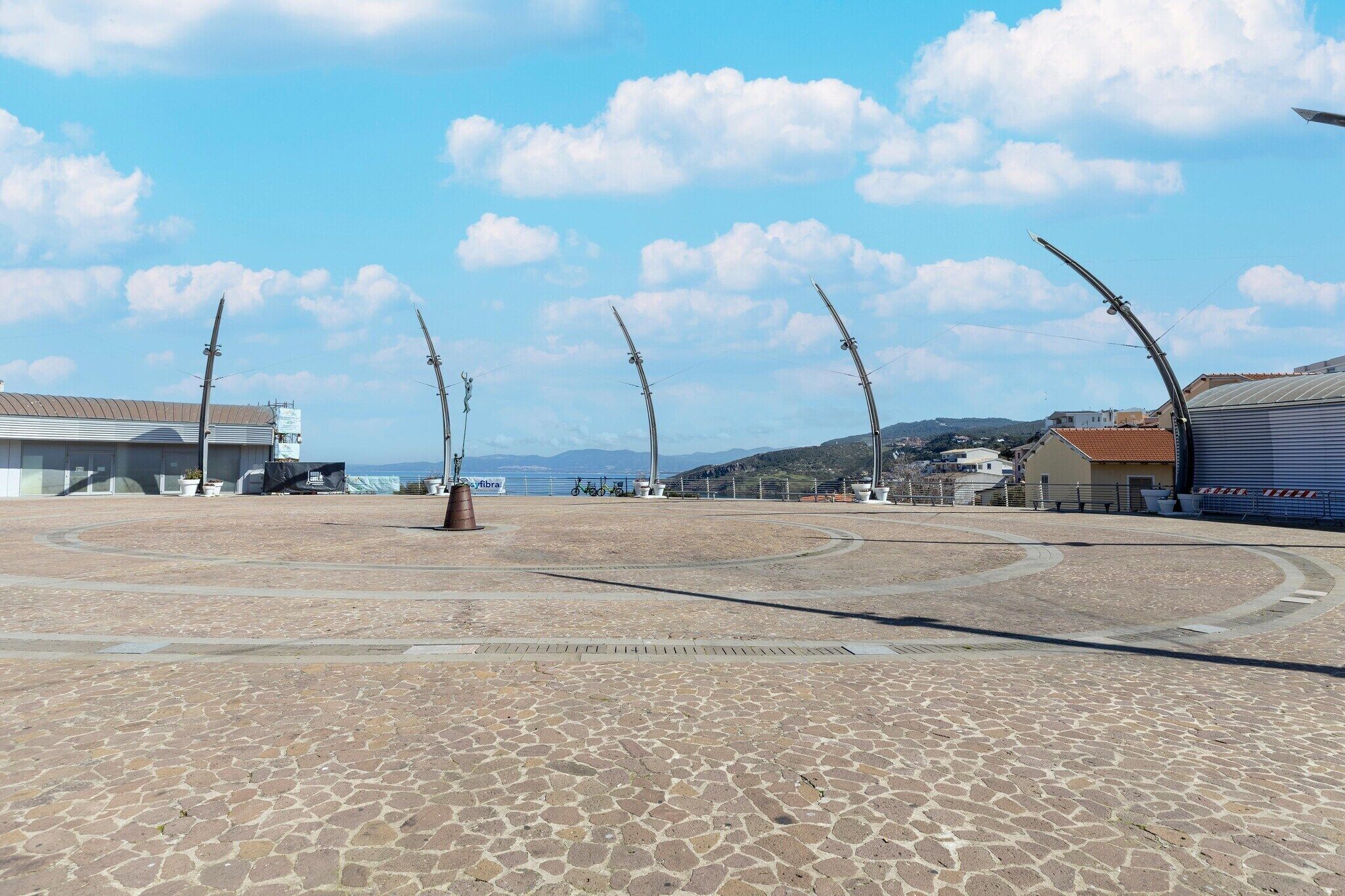 A paved plaza with curved metal structures, a building with a red roof, and a clear sky.