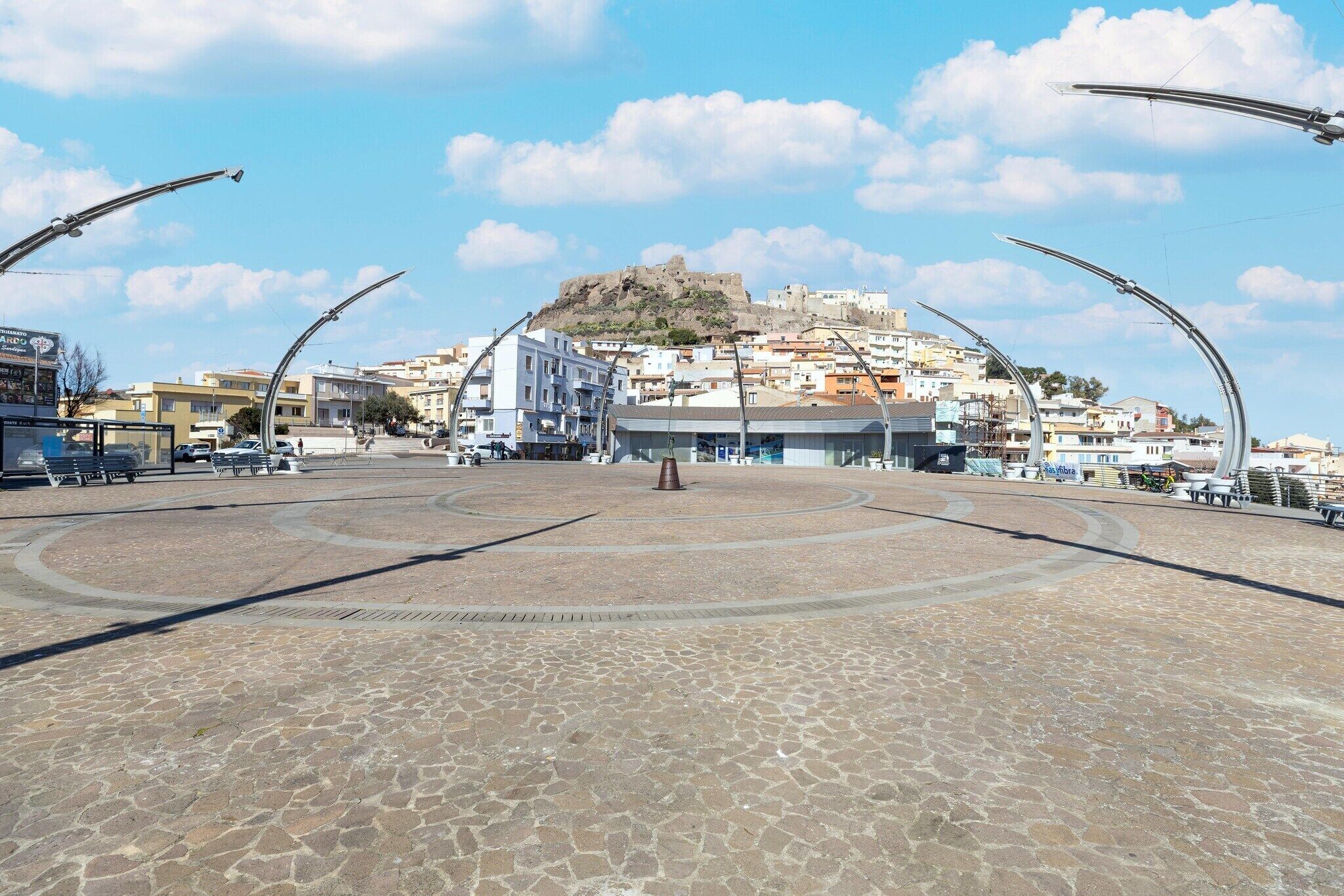 Ein gepflasterter Platz mit einem zentralen Denkmal, Gebäuden im Hintergrund und einem Hügel mit einer Festung.