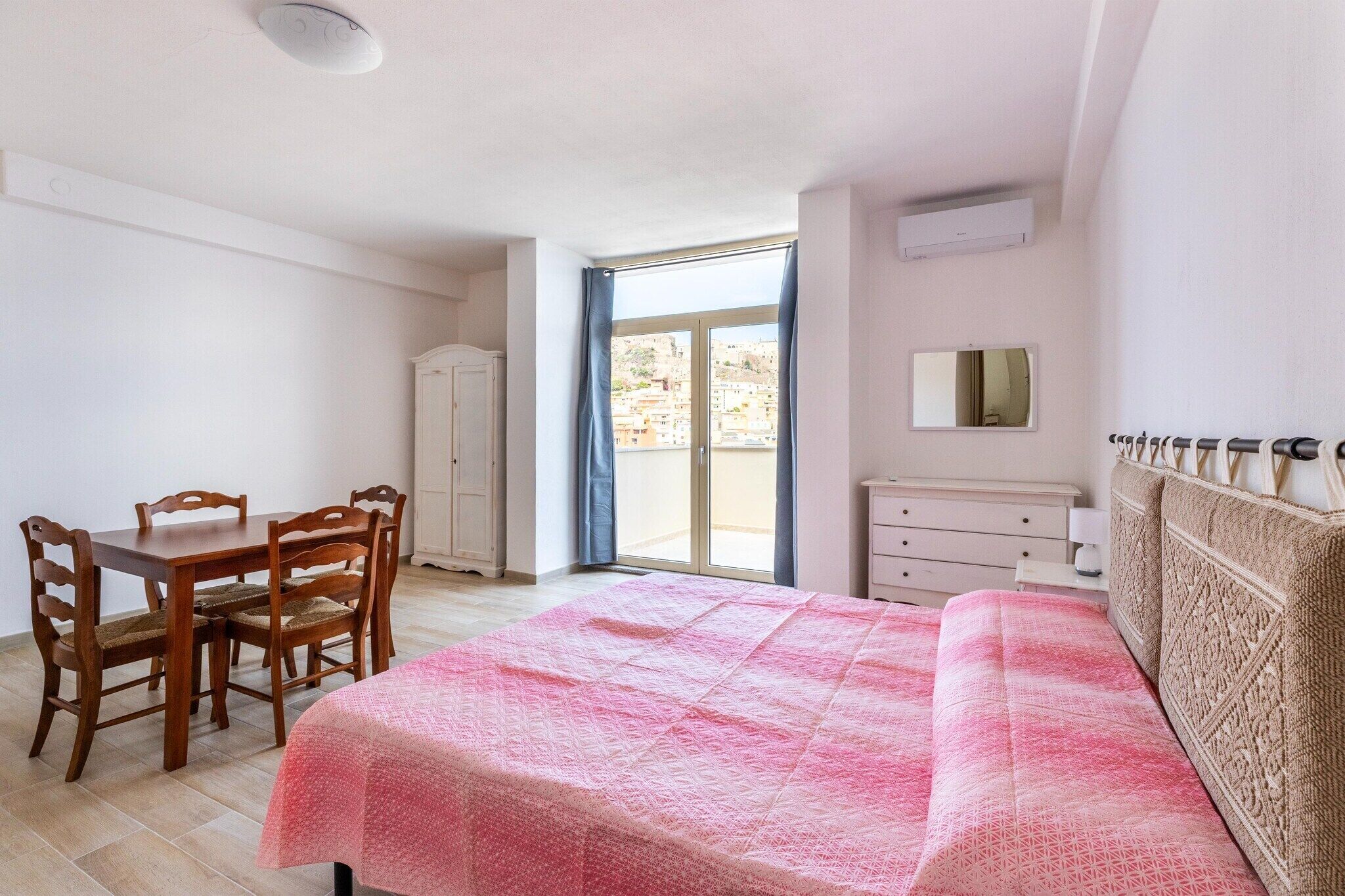 A bedroom with a bed, a desk with chairs, a dresser, and a sliding glass door to the outside.