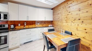 A modern kitchen with white cabinets, a wooden wall, a dining table with chairs, and a microwave. - Okanagan - Ponderosa Inn 408 - Hilltop Hideaway (Big White Mountain)