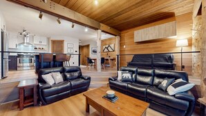 Un espacio habitable amplio con techo de madera, un juego de sofá de cuero negro, una mesa de comedor y una cocina al fondo.