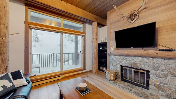 Una sala de estar acogedora con chimenea de piedra, un televisor de pantalla plana montado encima y un ventanal con vistas a una terraza nevada.