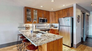 A modern kitchen with wooden cabinets, stainless steel appliances, a granite countertop, and bar stools. - Okanagan - Raven 104 (Big White Mountain)