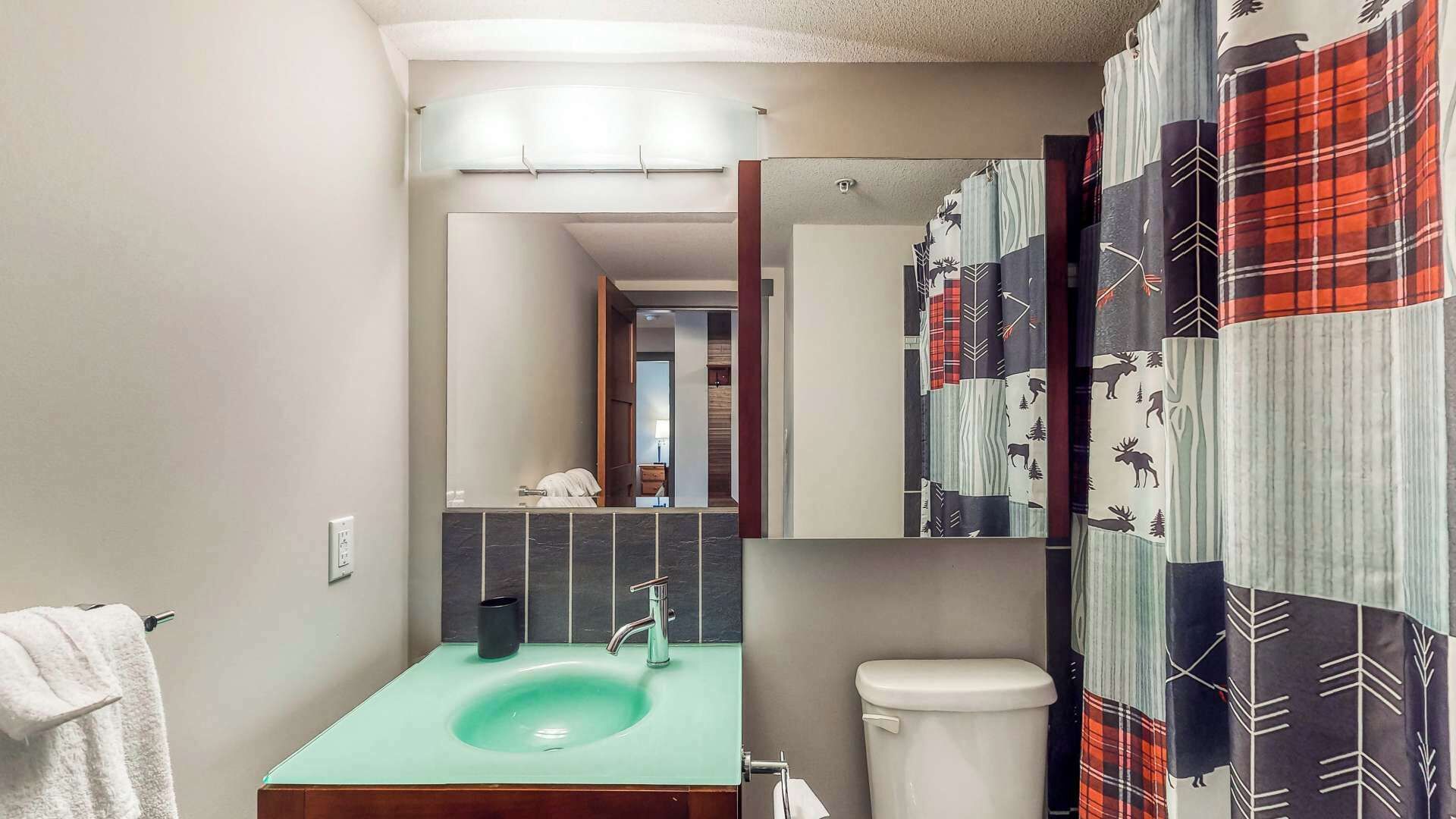 Une salle de bain avec un lavabo vert, des toilettes blanches, un miroir et un rideau de douche orné d’un motif de forêt.
