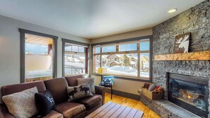 A cozy living room with a stone fireplace, leather sofa, and large windows overlooking a snowy landscape. - Okanagan - Raven 104 (Big White Mountain)