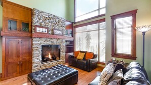A cozy living room with a stone fireplace, wooden cabinets, a leather sofa, and large windows. - Okanagan - Powder View 5 (Beaverdell)