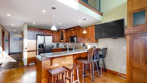 A modern kitchen with wooden cabinets, a stainless steel refrigerator, a microwave, and a large island with bar stools. - Okanagan - Powder View 5 (Beaverdell)