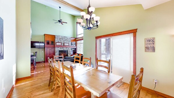 A dining area with a wooden table and chairs, a chandelier, and a stone fireplace. - Okanagan - Powder View 5 (Beaverdell)