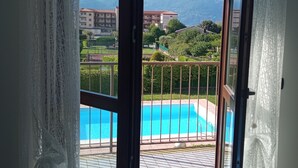 A room with a large sliding glass door leading to a balcony with a swimming pool, a view of mountains, and a building in the background. - Belmonte Holiday Apartment (Brezzo di Bedero)