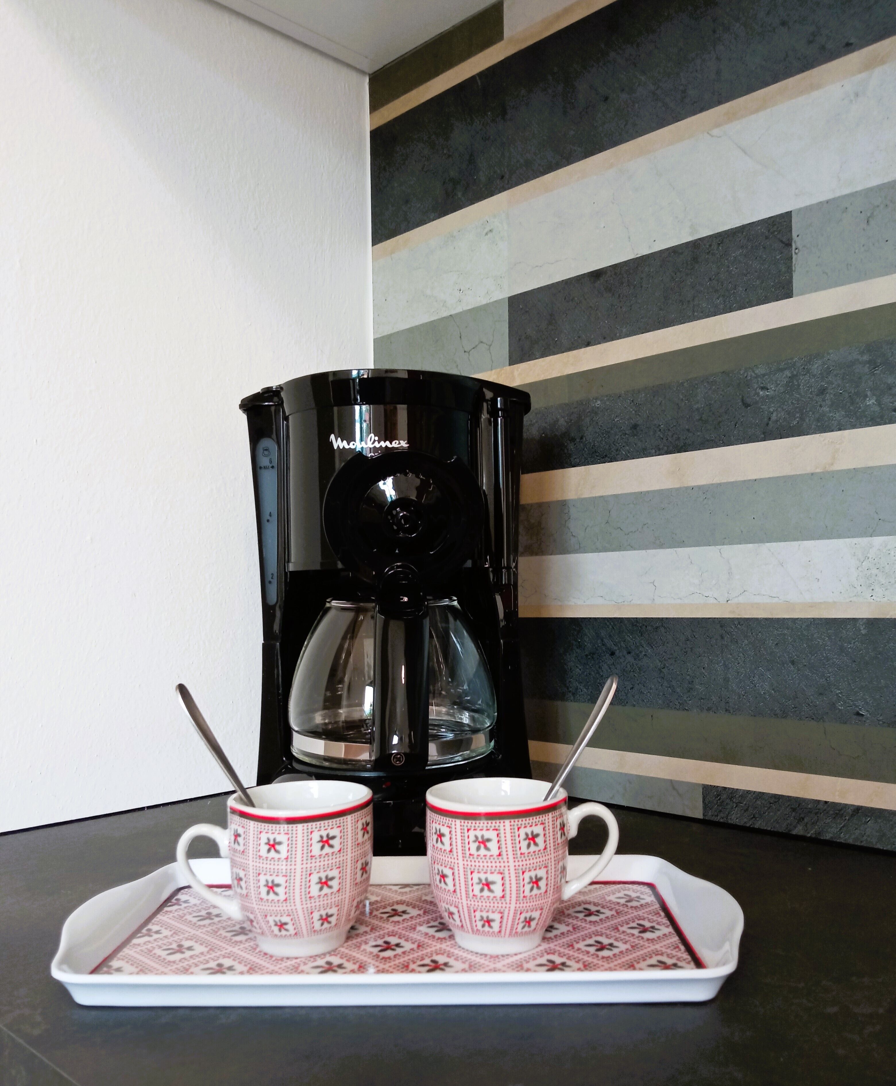 A black coffee maker on a tray with two patterned cups and spoons.