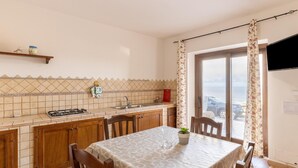 Cocina con estufa a gas, lavamanos y armarios de madera. Se observa una mesa de comedor con sillas y una planta en maceta. Se ve una ventana con cortinas, y un coche estacionado afuera.