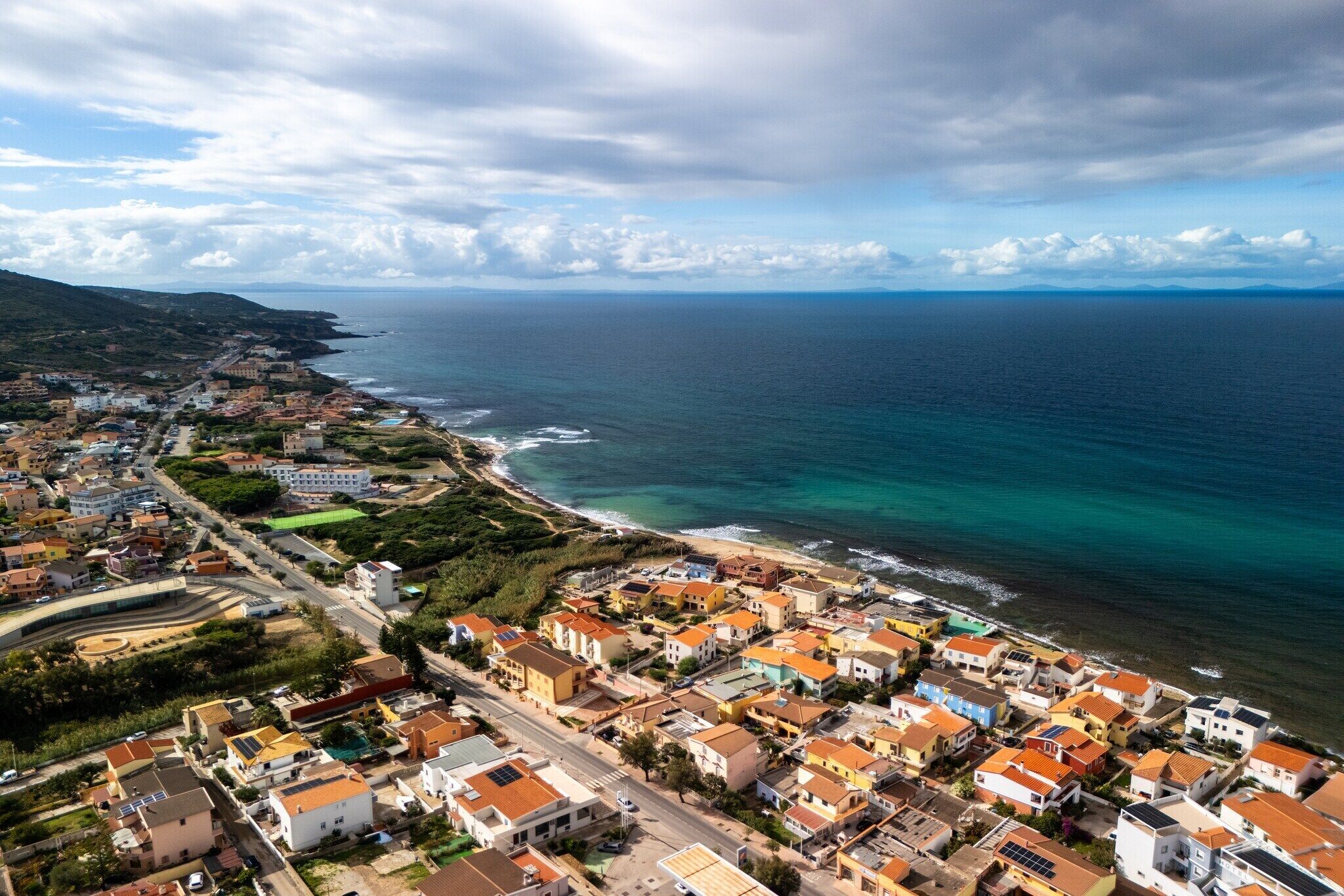 An aerial view of a coastal town with buildings along the shoreline, a clear view of the sea, and a hilly landscape in the background.