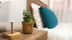 Una mesita de noche con una lámpara, una cesta tejida con una suculenta y una almohada azul turquesa sobre una cama con ropa de cama blanca.