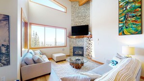A spacious living room with a stone fireplace, a large window with a view of trees, and a modern TV mounted on the wall. - Okanagan - Snowy Creek 10 (Beaverdell)