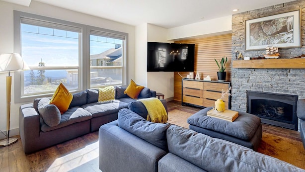 Ein modernes Wohnzimmer mit einem Ecksofa, einem Flachbildfernseher, einem Kamin und einem großen Fenster mit Blick ins Freie.