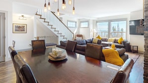 Ein geräumiger Wohnbereich mit einem dunklen Holzesstisch, Ledersesseln und einem großen Sofa mit gelben Zierkissen. Eine Treppe mit Holzgeländer führt auf eine obere Ebene.
