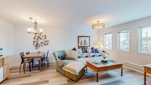 A cozy living room with a wooden dining table, comfortable seating, a decorative wall piece, and a large window with a view of snow-covered trees. - Okanagan - SnowCrest Village, Suite 5B (Beaverdell)