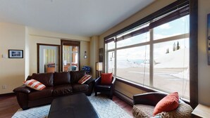 A living room with a brown leather sofa, a small armchair, and a bench. There is a large window with a view of a snowy landscape. - Okanagan - The Crescent 104 (Beaverdell)