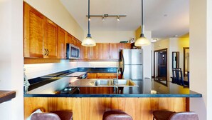 A modern kitchen with wooden cabinets, a stainless steel refrigerator, a microwave, and a large island with bar stools. - Okanagan - The Crescent 104 (Beaverdell)